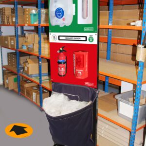 A safety station in a warehouse with shelves behind it. The station includes a mask and glove dispenser on top and a fire safety section with an extinguisher and alarm below. A bin with transparent bags is underneath. A yellow arrow is on the floor.