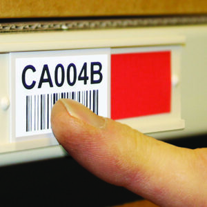 A finger pointing at a white label with a barcode and the text CA004B on a shelf. A red square is next to the label. The background includes a blurred cardboard box.