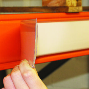 A hand is expertly placing a transparent label holder on an orange metal beam, part of innovative shelving or racking in a warehouse. This detail-oriented action reflects efforts to improve warehouse designs. A wooden pallet rests securely on top of the beam.