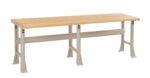 A long, rectangular wooden workbench with a smooth surface and metal legs. The bench has a natural wood finish and beige-colored legs, suitable for workshops or garages.