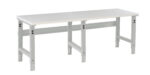 A long, rectangular white workbench with adjustable metal legs and support beams. The bench top has a smooth surface, and the legs have holes for height adjustment. The design is simple and functional, suitable for a workshop or garage.