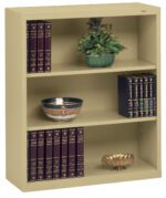 A beige bookshelf with three shelves. The top shelf holds a green plant in a pot. The middle shelf has a decorative bowl and several books. The bottom shelf contains a set of burgundy books and a small ornate bowl.