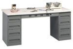 A gray workbench with multiple drawers, featuring a digital multimeter, circuit board, tools, and electronic equipment on top. The workbench has a smooth surface and power outlets underneath.