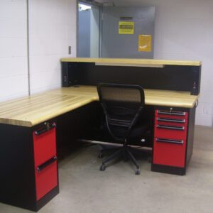 A corner desk with a wooden top and black base, featuring red drawers, is set up in a room with white walls. A black mesh office chair is positioned at the desk, and an open door in the background reveals another room.