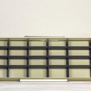 An empty, beige metal shelving unit with multiple compartments and black dividers sits against a plain white background. A single white tube lies in front of the shelving.