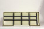 Empty beige and black storage shelf unit, placed horizontally against a plain white background. A white cylindrical rod lies at the base of the shelf.