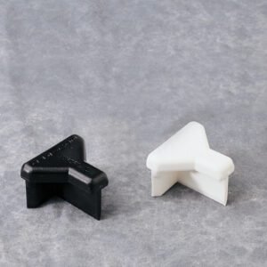 A black and a white plastic corner protector, shaped like a right angle, are placed on a light gray textured surface. They are designed to protect furniture or walls from damage.