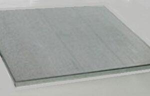 Image of a single sheet of transparent or frosted glass resting on a flat, light-colored surface. The glass has a subtle texture and reflects light slightly.