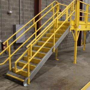 A yellow metal staircase with side railings leads to an elevated platform, forming part of a custom catwalk system in an industrial setting. The area features concrete walls and floor, with a fire extinguisher prominently displayed on the wall.