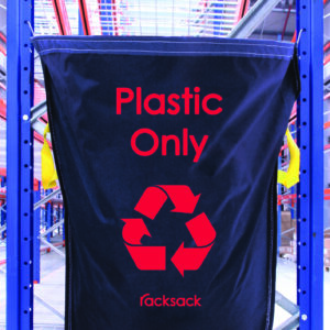 A black MH-USA Racksack - Plastic Only bag, featuring a red recycling symbol, hangs on a blue metal rack in an industrial space. The Racksack brand is prominently displayed with steel shelves and a ceiling grid in the backdrop.