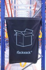 A large black MH-USA Racksack Symbol Only - Cardboard with a blue outline and bold racksack print is attached to blue metal shelves in a warehouse, hanging amidst orange and blue shelving units. In the background, cardboard boxes are neatly stacked.