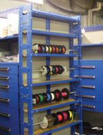 A blue metal shelf in a workshop holds various spools of colorful wire. The shelf is next to blue cabinets with multiple drawers. The background shows industrial piping and additional shelving.