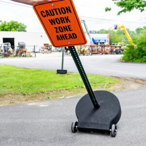 A hand holds a vibrant orange Caution Work Zone Ahead sign on a TCT Heavy Duty 60lb base with 1/2 steel axle and rubber wheels, sturdily placed on a paved path. Industrial equipment and lush greenery form the backdrop.
