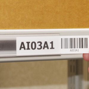 A hand applies a label marked AI03A1 and a barcode into an MH-USA Angled self-adhesive Ticket Holder 1.54 in. x 3.94 in. Pk 100 on a shelf, with a cardboard box visible in the background.