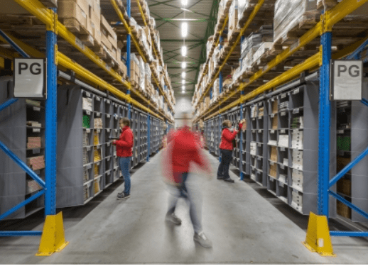 A blurred image captures a person walking briskly down an aisle in a massive warehouse. Shelves on both sides brim with boxes, while two people in red shirts, true storeganizers, efficiently pick items from the shelves. Overhead lights cast a bright glow across the scene.