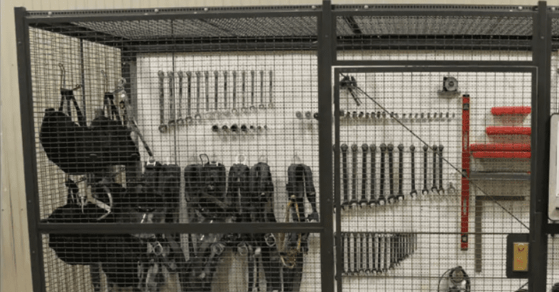 A storage area with various tools hanging on a pegboard reveals wrenches of different sizes neatly arranged. Black equipment hangs in the corner, and there are some red items on shelves to the right. The area is secured by a wire mesh gate, creating a makeshift security cage.