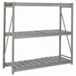 A large, empty industrial metal shelving unit with three levels and open sides. The shelves are made of parallel slats, and the unit is supported by a sturdy frame with cross braces. The color is a muted gray.