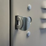 Close-up of a metallic lock hinge on a gray locker door. The hinge is partially open and surrounded by round rivets, with the locker surface featuring a subtle vertical pattern.
