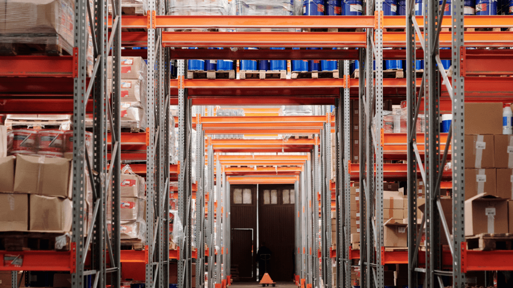 The warehouse interior boasts cost-effective storage solutions with tall, orange, metal-framed shelves filled with boxes. These versatile Borroughs Rivet-Span Shelving units extend down a long aisle. A cone is positioned on the floor near a closed doorway at the end of the aisle.