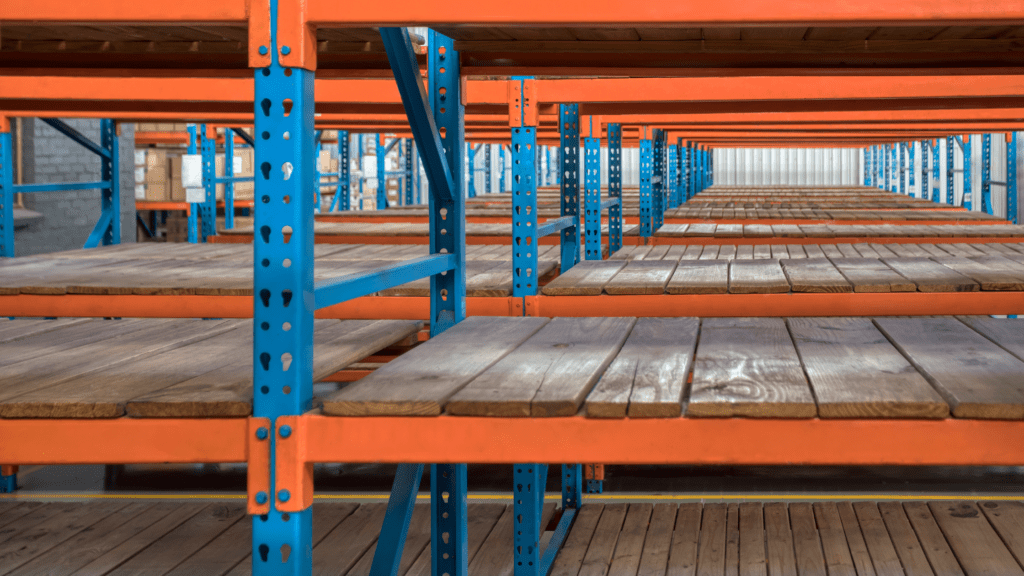 Empty industrial warehouse shelving features blue metal supports and orange frames, showcasing an impressive Storage Solution. Wooden shelves extend horizontally, creating repetitive lines and patterns. The Borroughs Rivet-Span Shelving ensures the space appears spacious and well-lit.