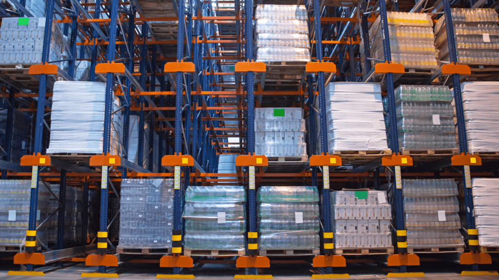 The warehouse interior features tall, blue and orange racks filled with stacked pallets, showcasing efficient storage solutions. Pallets, wrapped in clear plastic, hold various boxed and bottled products. The well-organized space benefits from bright overhead lighting.