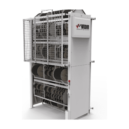 White vertical storage unit with multiple rotating shelves holding rolls of material, showcasing advanced automated storage solutions. A safety cage surrounds the shelves, and a control panel is visible on the side. The word VIDIR is printed in red and black on the front panel.