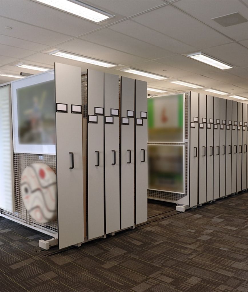 Storage room with GSA Mobile Shelving features vertical sliding racks displaying large framed paintings. The racks have black handles and labels at the top. The carpeted floor and recessed lighting complement the space. Some artwork is visible, though blurred, ensuring efficient inventory organization.