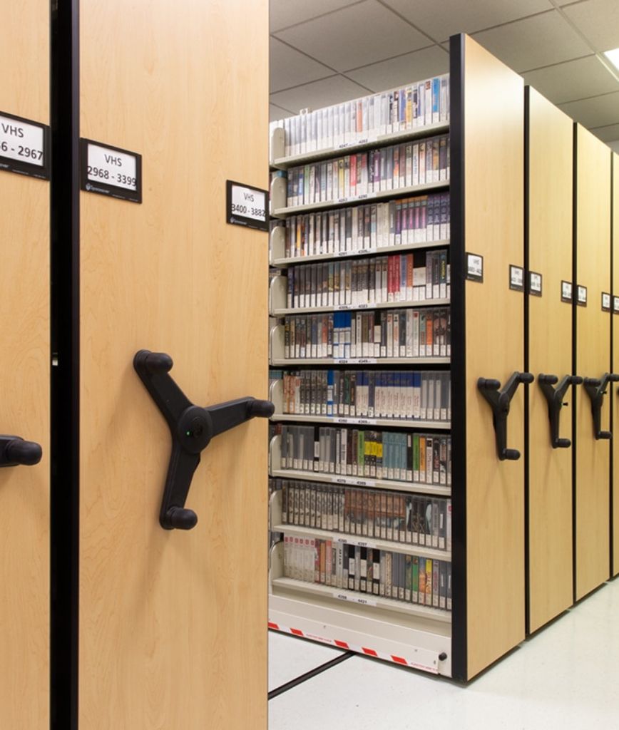A storage system with mobile shelving, designed for efficient inventory organization, holds rows of VHS tapes. Each shelf features a numbered label. Large black wheels are attached to the wooden panels for easy movement, embodying space management solutions in a well-lit area with white flooring.
