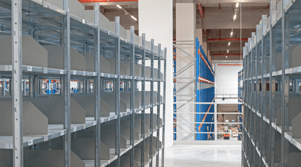 Empty metal shelves in a well-lit warehouse, with a view of the floor below. This efficient layout highlights how catwalk shelving systems can improve your bottom line. The blue and orange shelving in the background underlines the benefits for industries utilizing such innovative storage solutions.