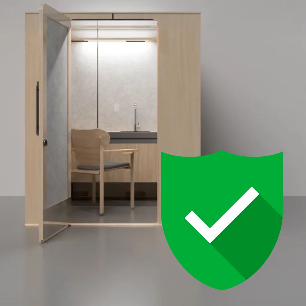 A wooden phone booth-like structure with a chair inside stands in a gray room. The booth has a glass door, reminiscent of Lactation Pods found in airports. In front, a large green shield icon with a white checkmark is prominently displayed, echoing the safety assured by the PUMP Act.