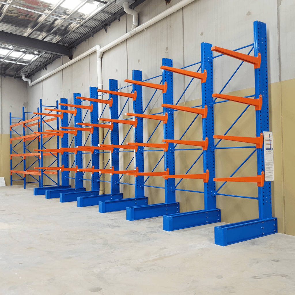 Blue and orange industrial steel shelving units line a warehouse wall with a concrete floor and high ceiling. Designed for material handling, these units are empty and evenly spaced, creating an efficient, clean, and organized appearance.