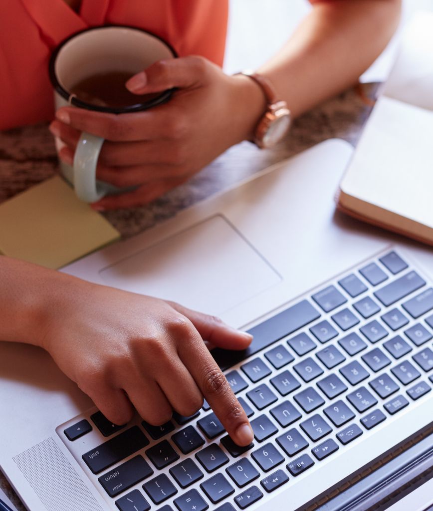 A person in an orange shirt and watch balances work with leisure, using a laptop one-handed while sipping from a mug. They sit at a desk, where convenient storage options neatly tuck away essentials, ensuring their workspace remains organized and efficient.