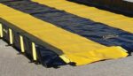 The yellow and black Justrite Track Runner, measuring 3X10, rests on concrete. Designed for outdoor use, it boasts sturdy edges and textured fabric. Small clips hold it securely in place, emphasizing its safety features.