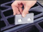 A hand displays a Justrite Stainless Steel Joining Clip, a rectangular metal piece with dual square cutouts, above a black grid-like surface, highlighting its precision and robust design for sturdy applications.