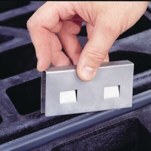 A hand displays a Justrite Stainless Steel Joining Clip, a rectangular metal piece with dual square cutouts, above a black grid-like surface, highlighting its precision and robust design for sturdy applications.