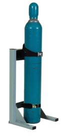 A tall, cylindrical blue gas cylinder is secured upright with two black straps in a gray Justrite Gcyl Stnd, 1Cyl, Lckpst, Stl. The cylinder has a rounded top and some visible scuff marks showing wear.