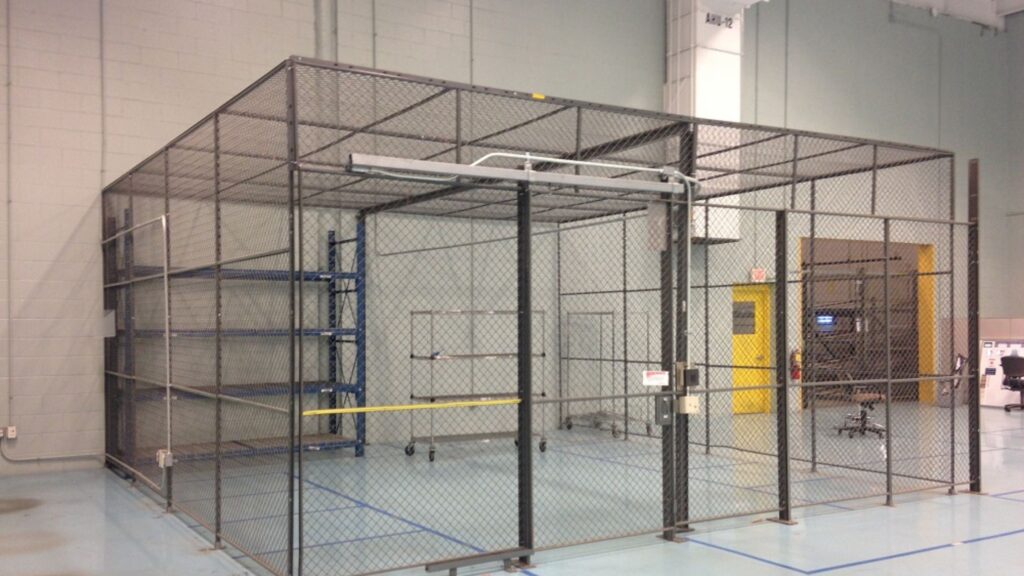 A large industrial cage made of chain-link fencing, tailored for theft prevention solutions, is situated inside a warehouse. It features shelves and racks with a sliding door marked by yellow caution tape. The floor is a light blue, emphasizing its role in enhanced warehouse security.