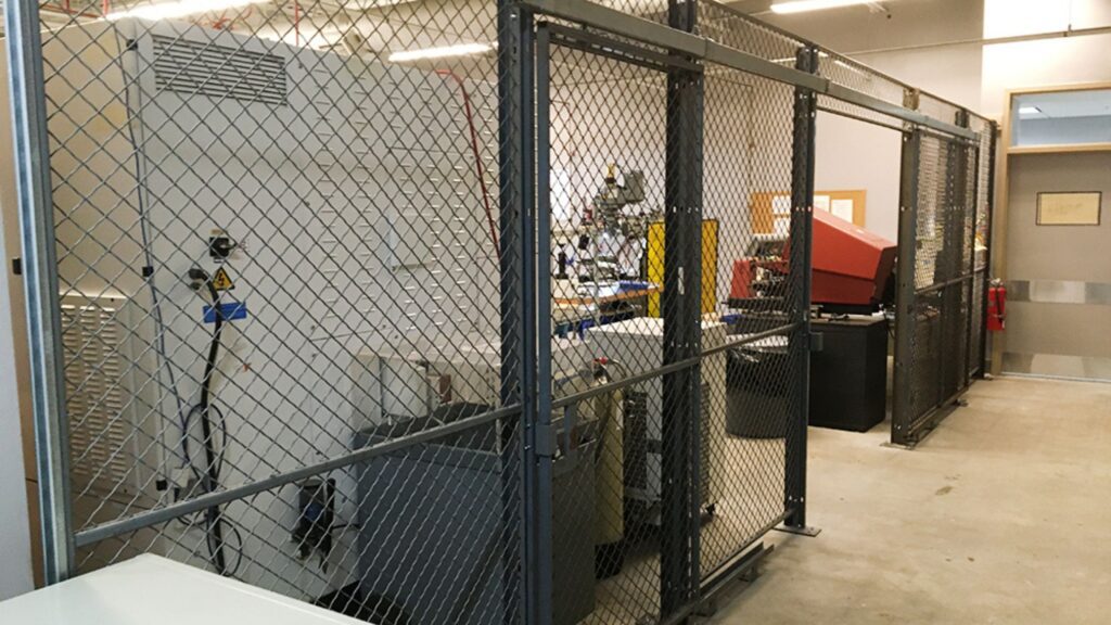 A secured industrial area is enclosed by a chain-link fence, offering asset protection. Inside are various large machines and equipment, including a white cabinet and a red machine. The surrounding concrete-floored workspace features overhead lighting for enhanced visibility.