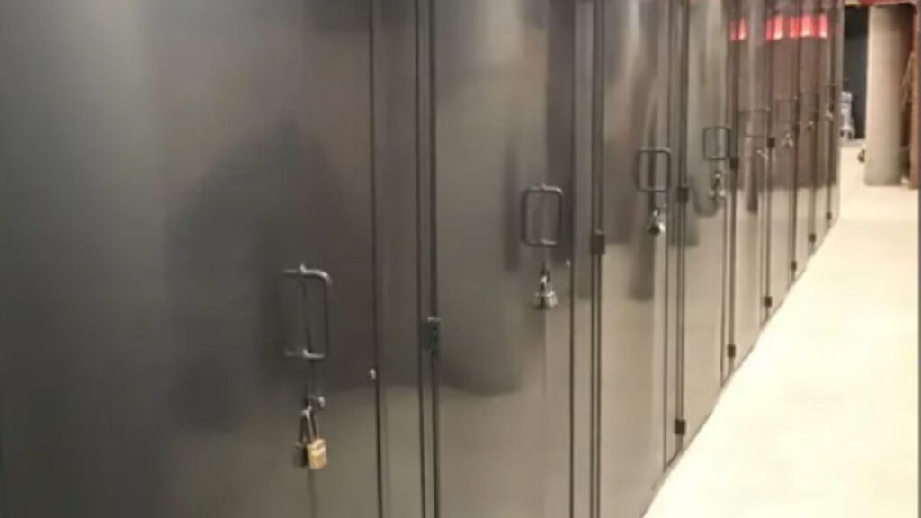 A row of dark gray metal lockers with padlocks on each door, embodying secure storage solutions, is lined up along a hallway. The lockers feature rectangular handles, and the floor is light-colored concrete. The corridor extends into the distance.