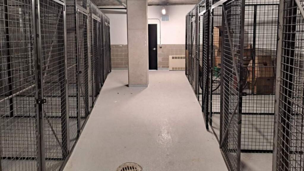 A hallway lined with metal storage cages and a concrete column. The floor is a light-colored smooth surface, and at the corridors end, a dark door with a security lock provides secure storage solutions. The area is illuminated by overhead lights, offering convenient storage options.