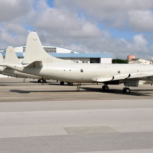 Three white military aircraft are parked on an airbase runway under a partly cloudy sky. Nearby, ballistic-rated modular buildings stand ready alongside guard shacks. The planes are lined up in a row, facing left, with hangars and other airport structures in the background.