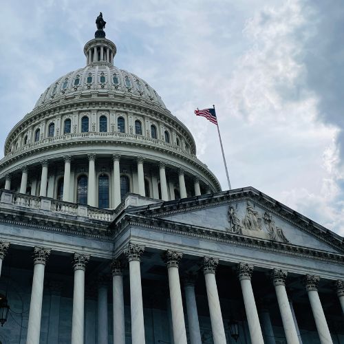 The image showcases the United States Capitol building with an American flag waving atop, set against a cloudy sky. The dome and detailed architecture stand out, resembling the strength and resilience of ballistic guard shacks with their durable design and protective structure.