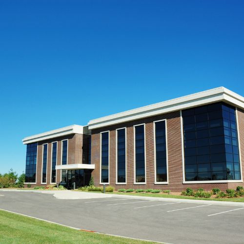 A modern, two-story office building with large glass windows and a brick facade features a guard shack built with ballistic rating. Surrounded by a well-maintained parking area and landscaping, it stands gracefully under a clear blue sky.