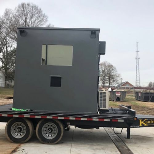 A large, gray rectangular structure with a window, resembling a mobile guard booth, is mounted on a flatbed trailer parked outdoors. Trees and a building form the backdrop. The trailer rests on a paved surface with grassy areas extending beyond it.