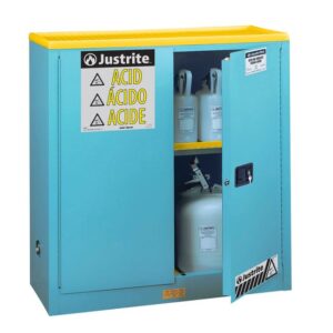 A blue Justrite Cabinet, model Acid Sfty 30G Man Blu, with a yellow top is partially open, revealing plastic containers inside. The door is labeled with caution signs and ACID in various languages for universal safety and clarity.