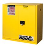 The yellow Justrite Cabinet, model Flam 30G, Sc/Bifld Yl, has two doors with a black handle on the right door secured by a padlock. Red and white FLAMMABLE KEEP FIRE AWAY labels are prominently displayed alongside caution signs near the base.