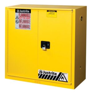 The yellow Justrite Cabinet, model Flam 30G, Sc/Bifld Yl, has two doors with a black handle on the right door secured by a padlock. Red and white FLAMMABLE KEEP FIRE AWAY labels are prominently displayed alongside caution signs near the base.