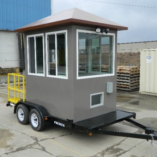 A small, portable gray guard shack on a trailer with wheels, offering flexible security structures. It features large windows and a brown roof, situated in an industrial area. The trailer includes a tow hitch and a yellow railing on one side.