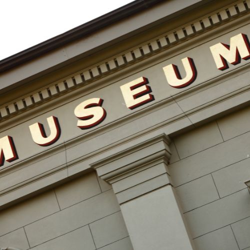A close-up of a buildings facade showcases the word MUSEUM in large, bold letters. The golden letters with red outlines stand out against classical architectural elements like columns and molding, seamlessly blending tradition with modern innovation, much like electric mobile shelving inside.