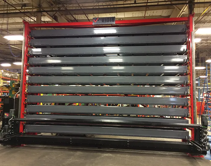 A Vidir Vertical Lift for Bar Stock Storage enhances the large industrial storage rack with multiple horizontal, stacked shelves. The gray metal shelves are secured in a red frame inside a bustling warehouse filled with machinery and equipment, boosting manufacturing efficiency.